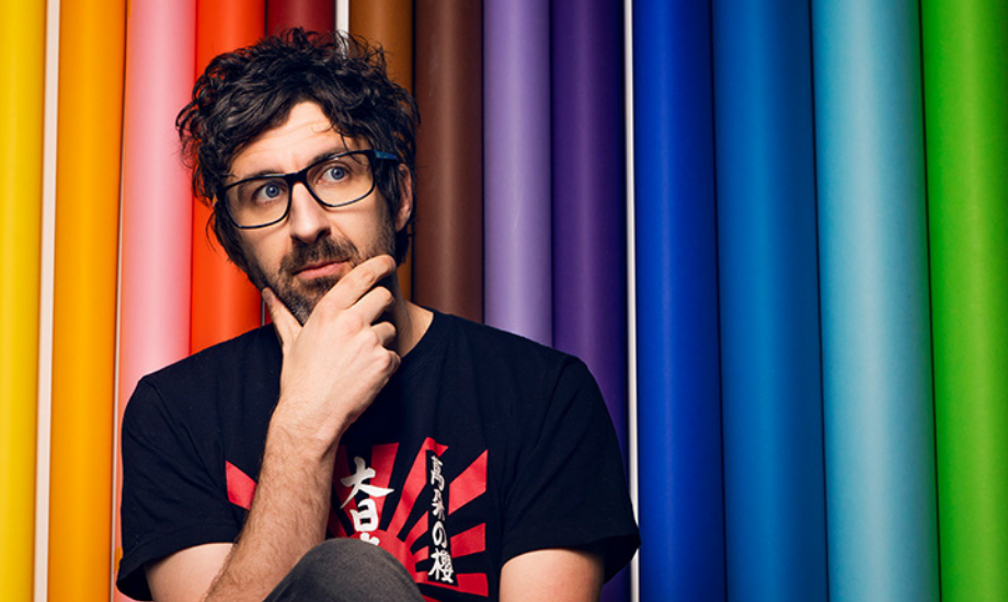 Mark Watson ponders in front of a rainbow background.